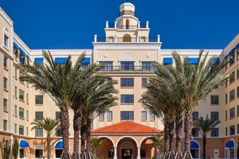 Front entrance of a multi-story Mediterranean-style building with an arched portico and rows of palm trees under a clear blue sky.
