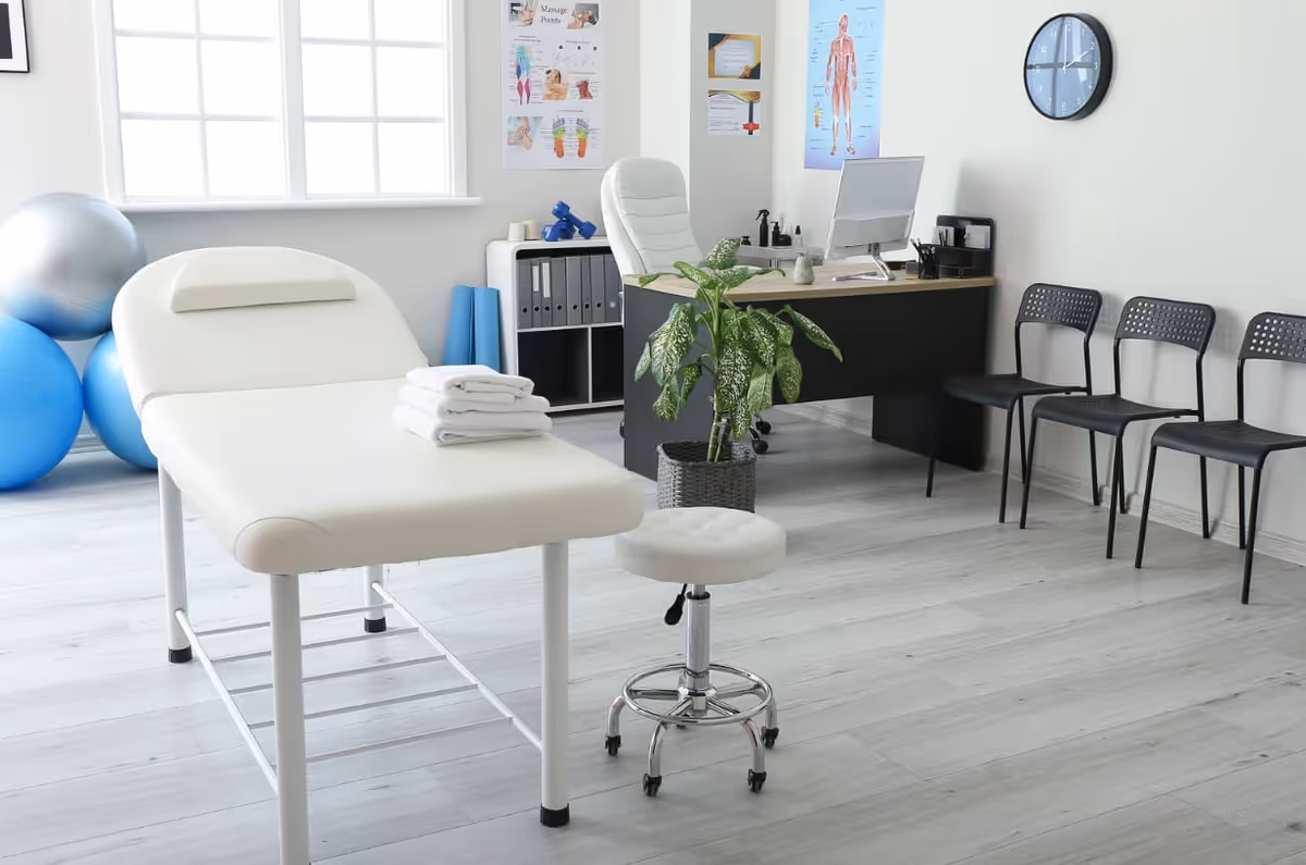 A clean medical treatment room with an examination table, desk, plant, and seating.