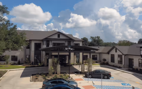 Front entrance of a two-story senior living building with a covered porte-cochère, parking spaces and cars under a partly cloudy sky.