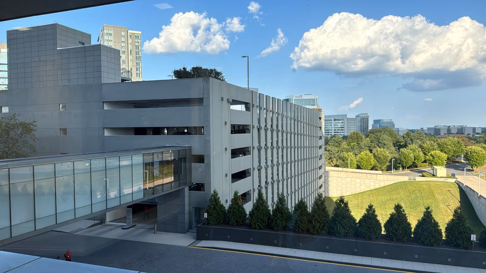 Modern multi-story parking garage with a glass walkway beside a landscaped, tree-lined area.