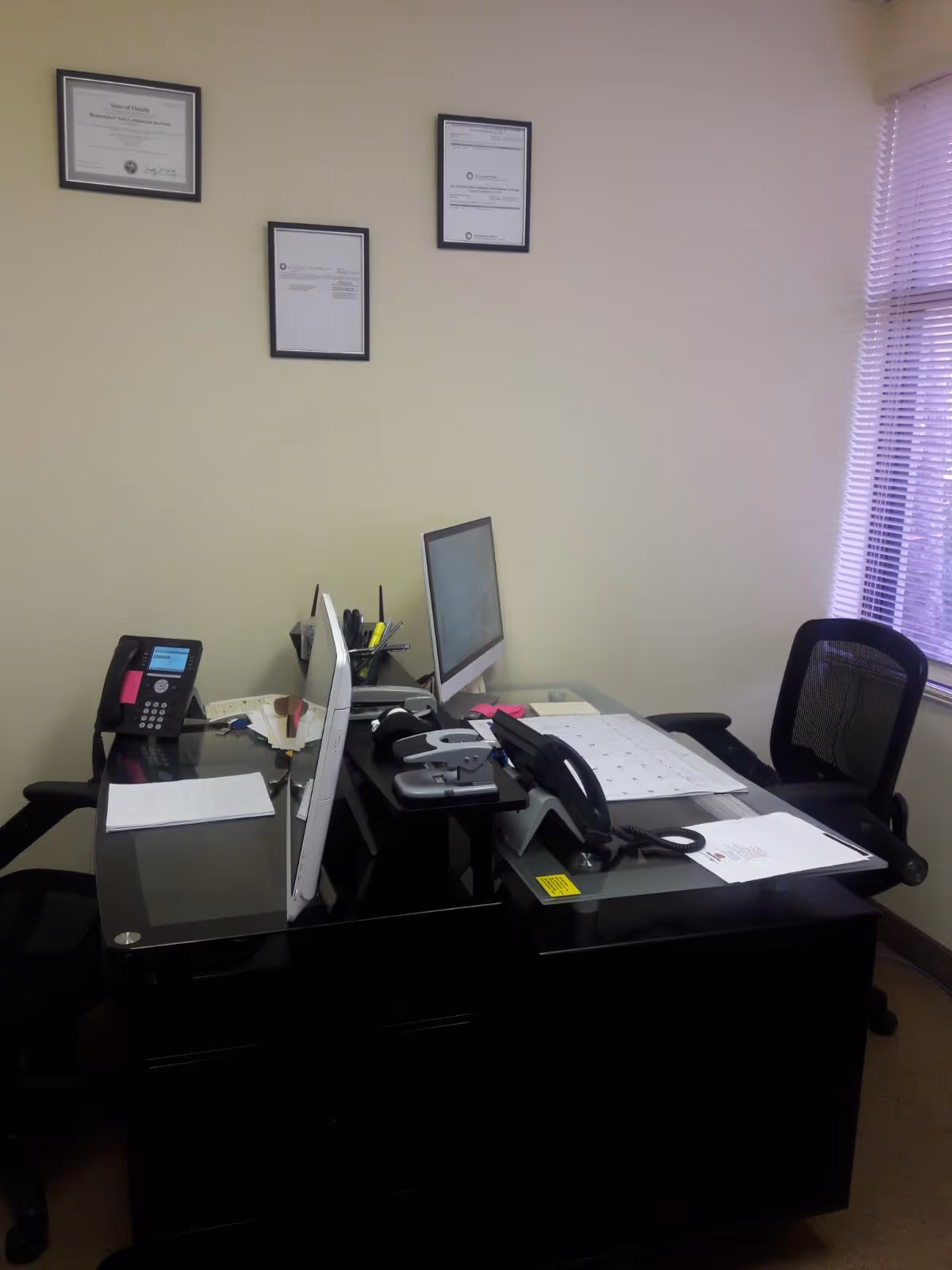 An office workspace with a glass desk, computer, phone, and scattered papers.