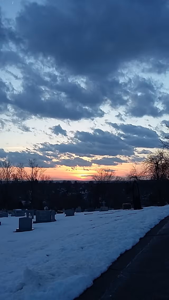 Snow-covered cemetery at sunset with leafless trees and headstones along a path.