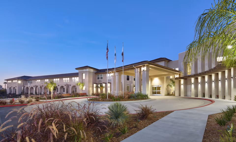 Front exterior of a senior living building with a covered porte-cochère, flagpoles, landscaped driveway and evening sky.