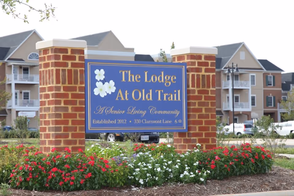 Entrance sign reading 'The Lodge At Old Trail' mounted between brick pillars with flowers and the senior living building behind it.