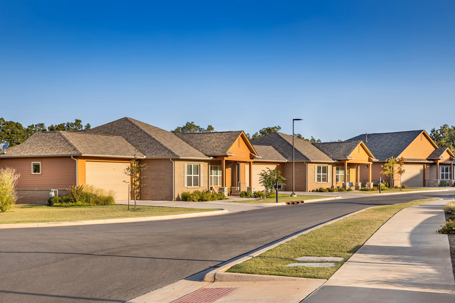 A row of single-story residential buildings with garages, set along a paved road with sidewalks and street lamps under a clear blue sky.