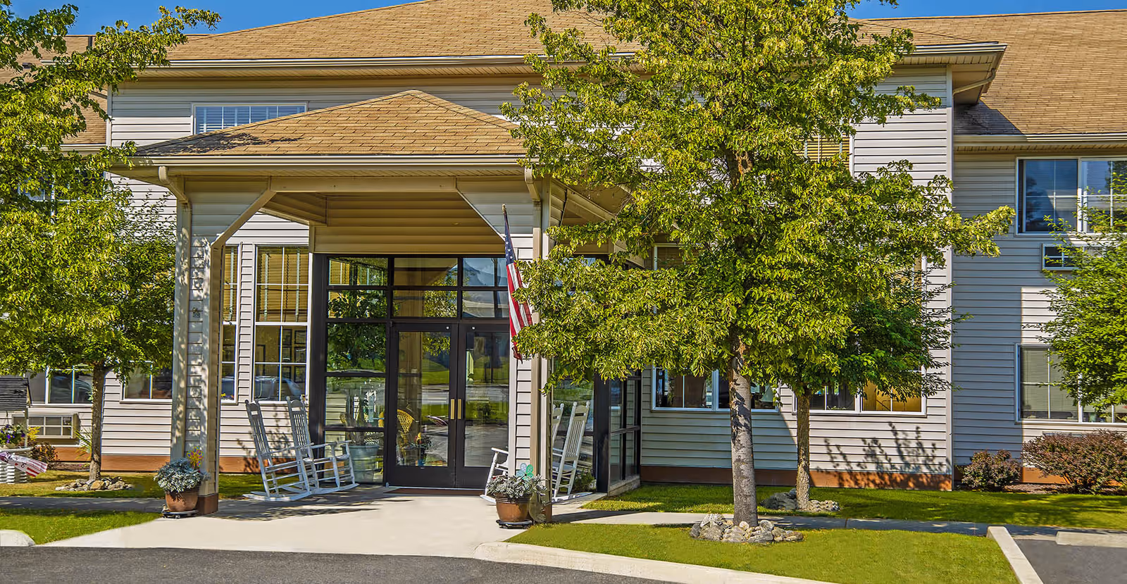 Covered main entrance of a senior living building with rocking chairs on the porch, an American flag, and trees in front.