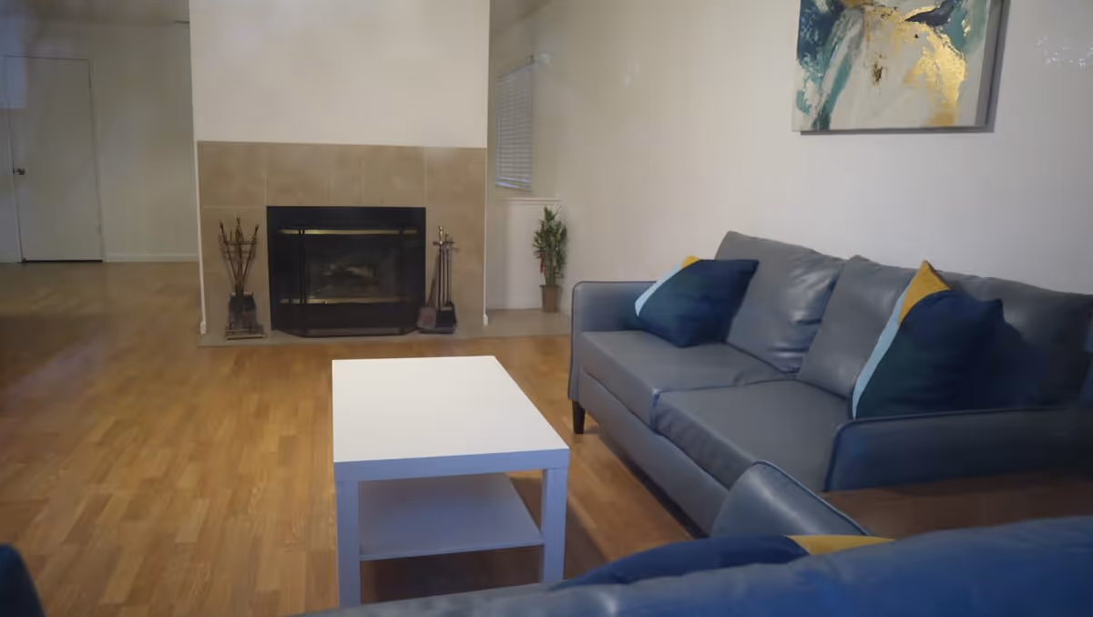 A living room with a gray leather sofa adorned with blue and yellow pillows, a white coffee table in front, a fireplace with beige tiles, a small plant in the corner, and a painting on the wall above the sofa.