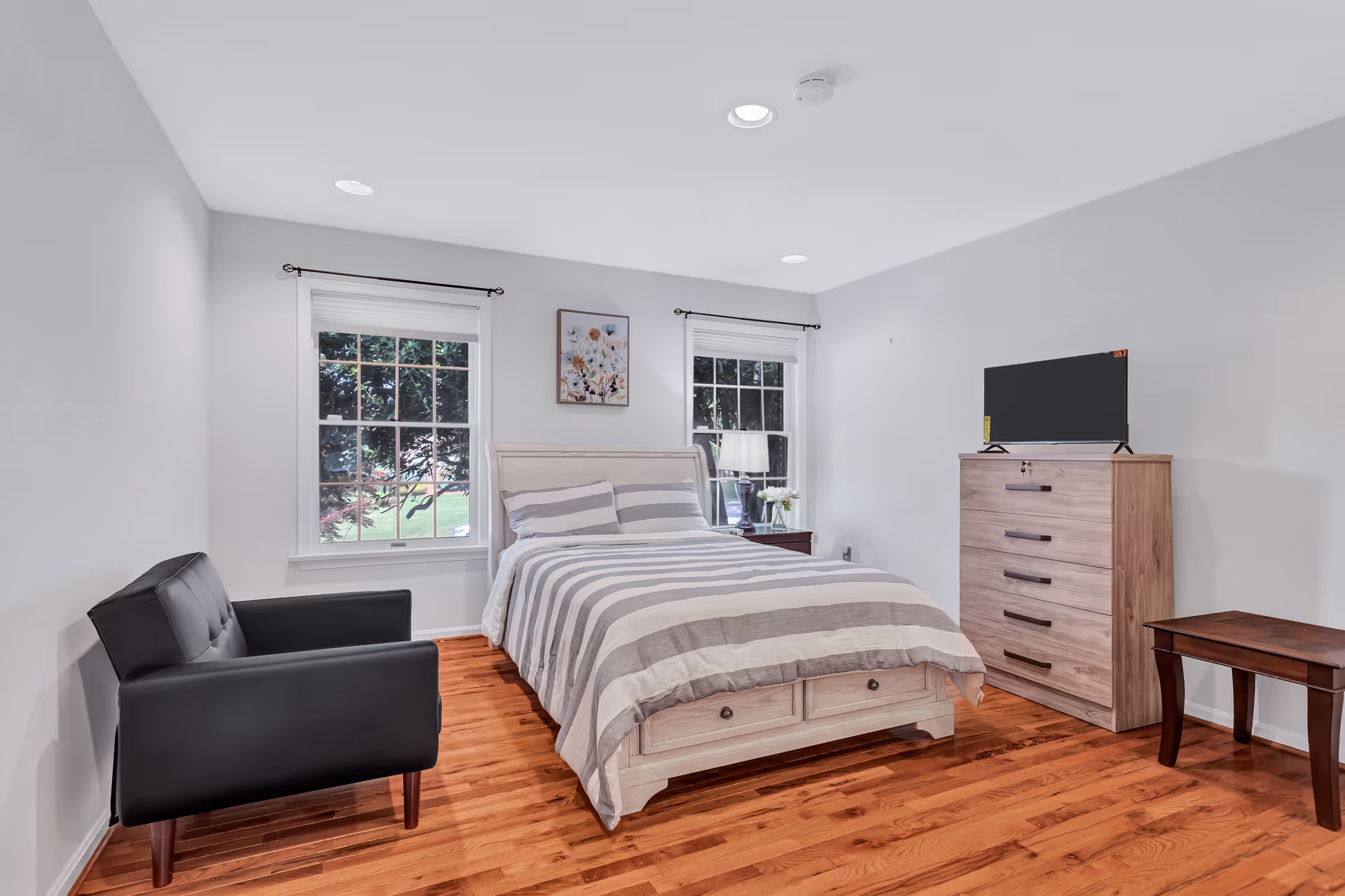 A furnished bedroom with a bed, dresser, TV, and black chair in a light-filled room.