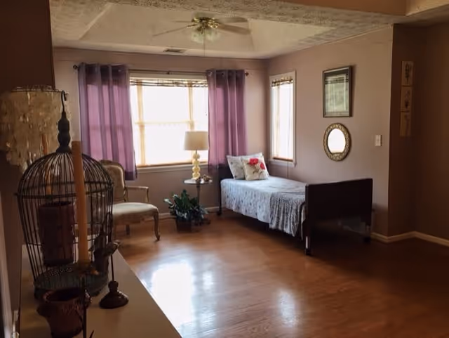 A cozy bedroom with a single bed covered in a light gray blanket and a decorative pillow. The room has wooden flooring, a window with purple curtains, a small side table with a lamp, a chair, and some wall decorations including a round mirror and framed pictures.