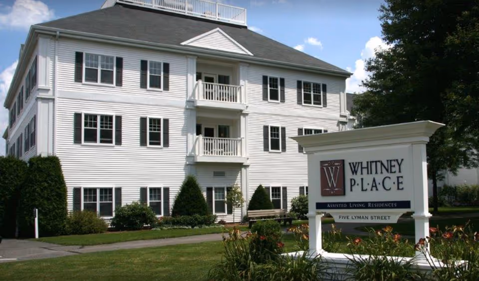 White three-story assisted living building with balconies and landscaped lawn and a large sign reading "WHITNEY PLACE" in front.