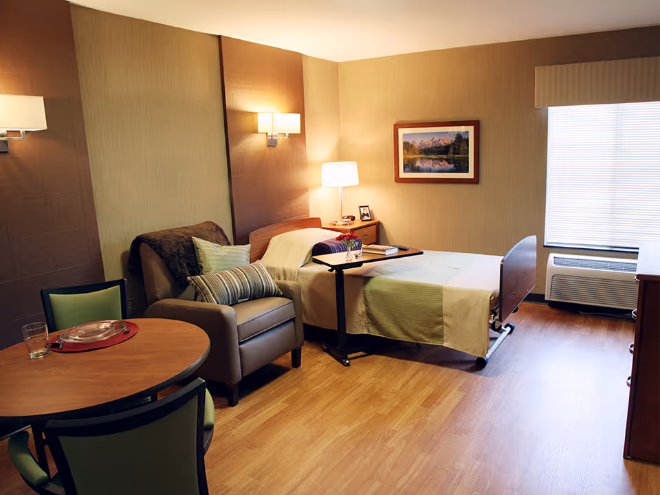 A cozy senior living room with a single bed, a bedside table with a lamp, a comfortable armchair with pillows, a round wooden table set with a plate and glass, and a window with blinds letting in natural light.