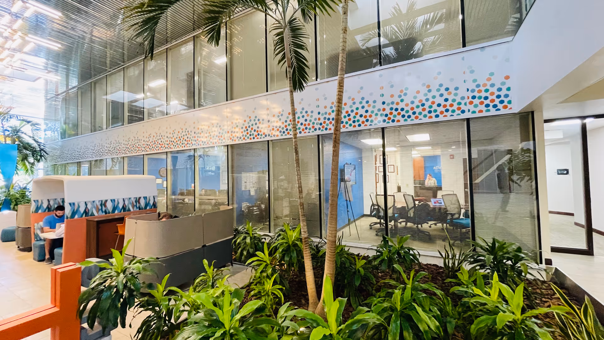 Indoor modern lobby with glass walls, tropical plants, and seating areas in a senior living facility.