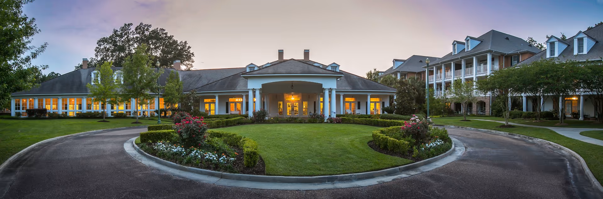 Panoramic front view of a senior living facility with a circular driveway, landscaped lawn, and a columned main entrance at dusk.