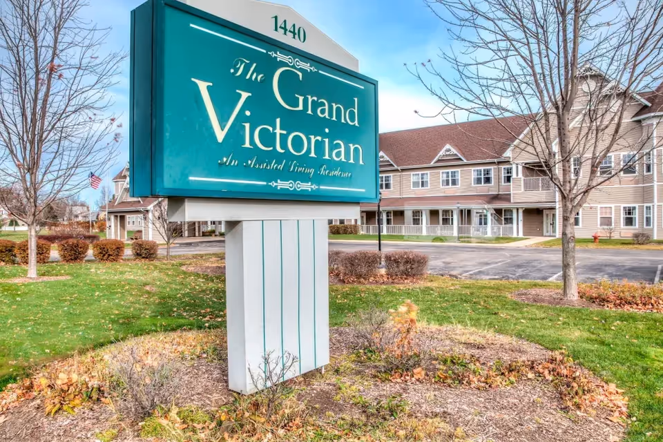 Exterior view of The Grand Victorian senior living facility with a large teal sign in Sycamore.