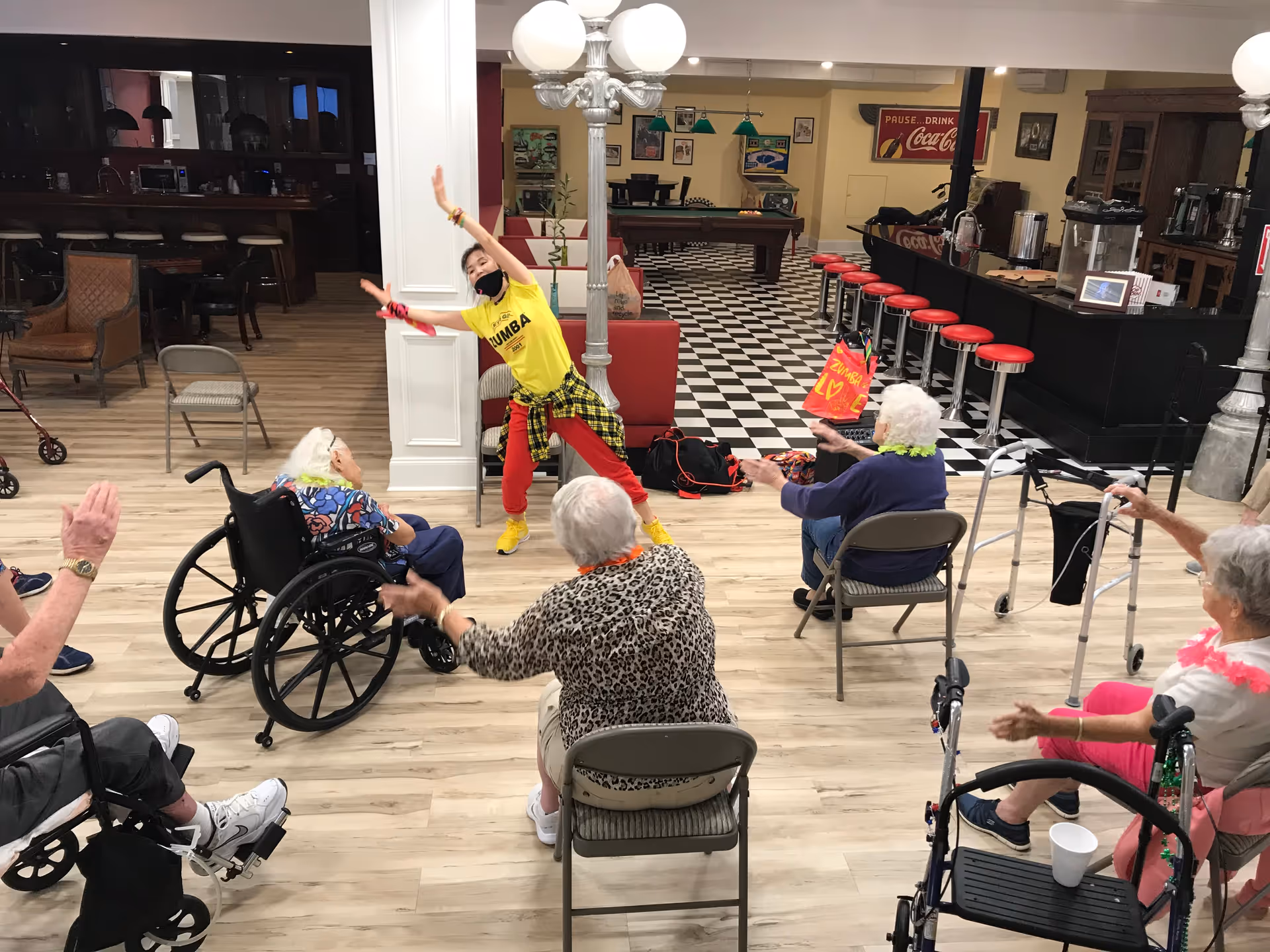 A group of elderly people seated in chairs and wheelchairs participate in a seated exercise class led by an instructor wearing a yellow Zumba shirt and red pants. The room has a wooden floor, a black and white checkered area with a pool table, and a bar with red stools in the background.