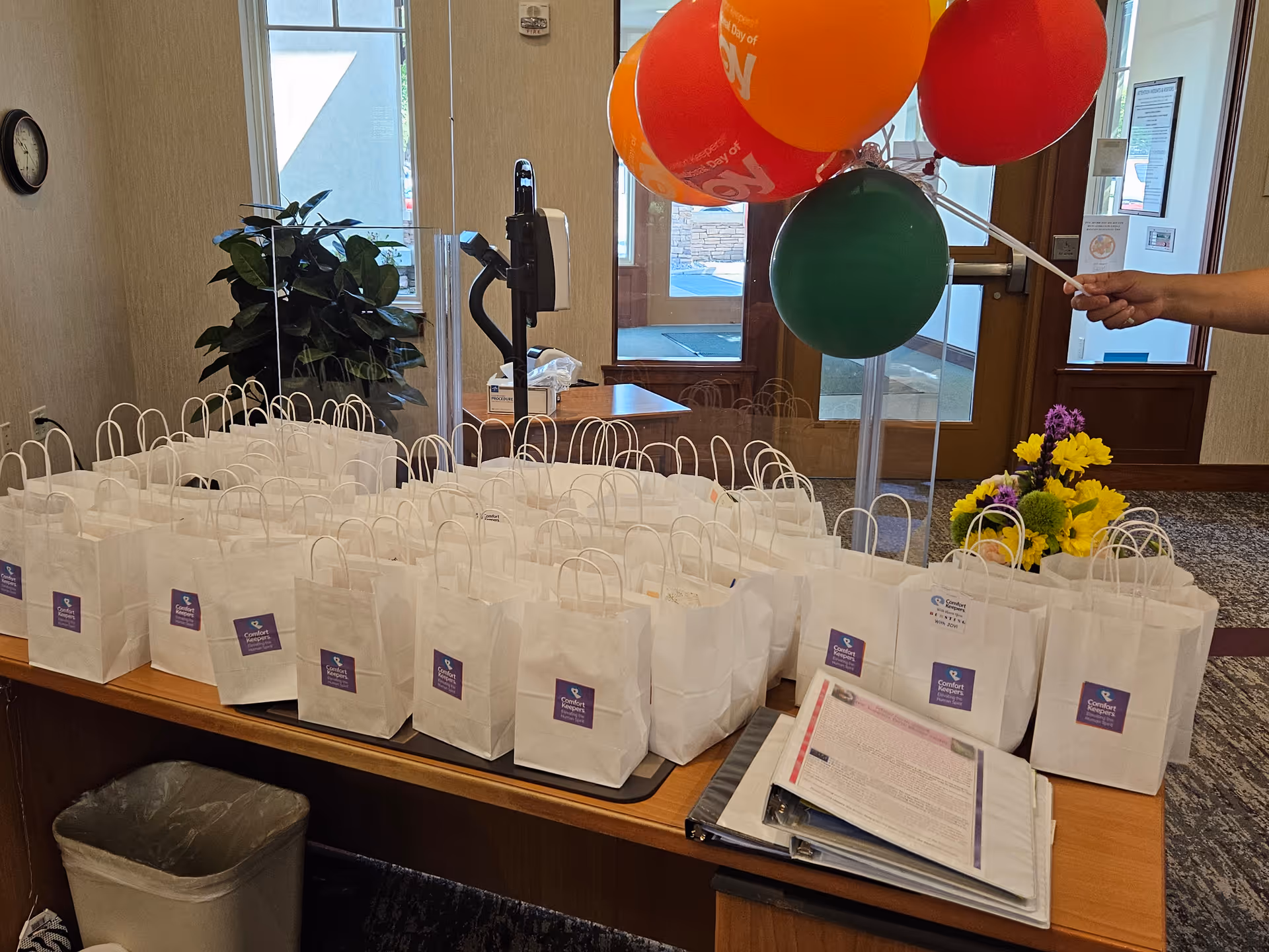 Lobby desk with white gift bags, balloons, and flowers in a care facility.