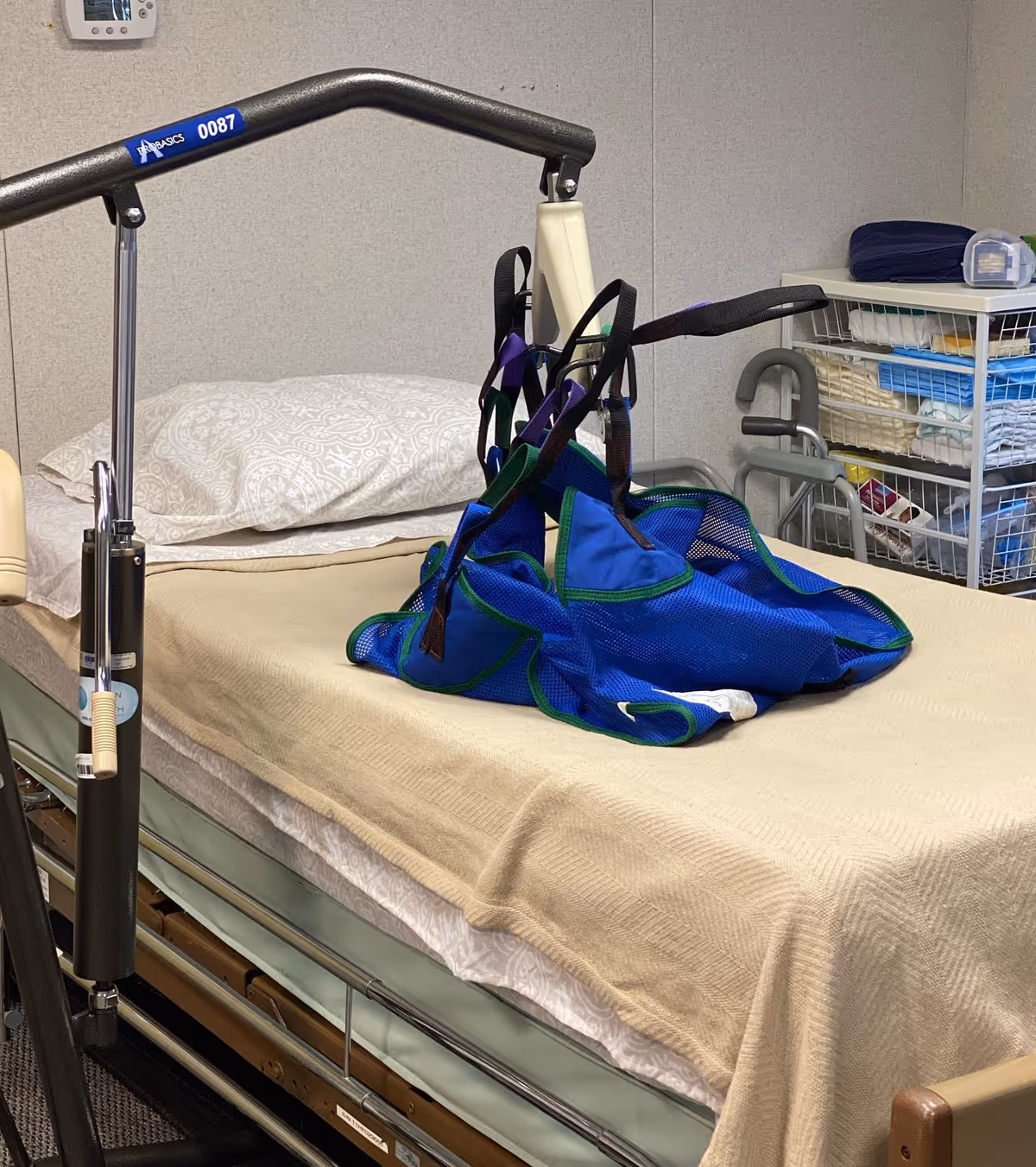 Hospital bed with trapeze frame, blue patient sling, and medical supplies in a room.