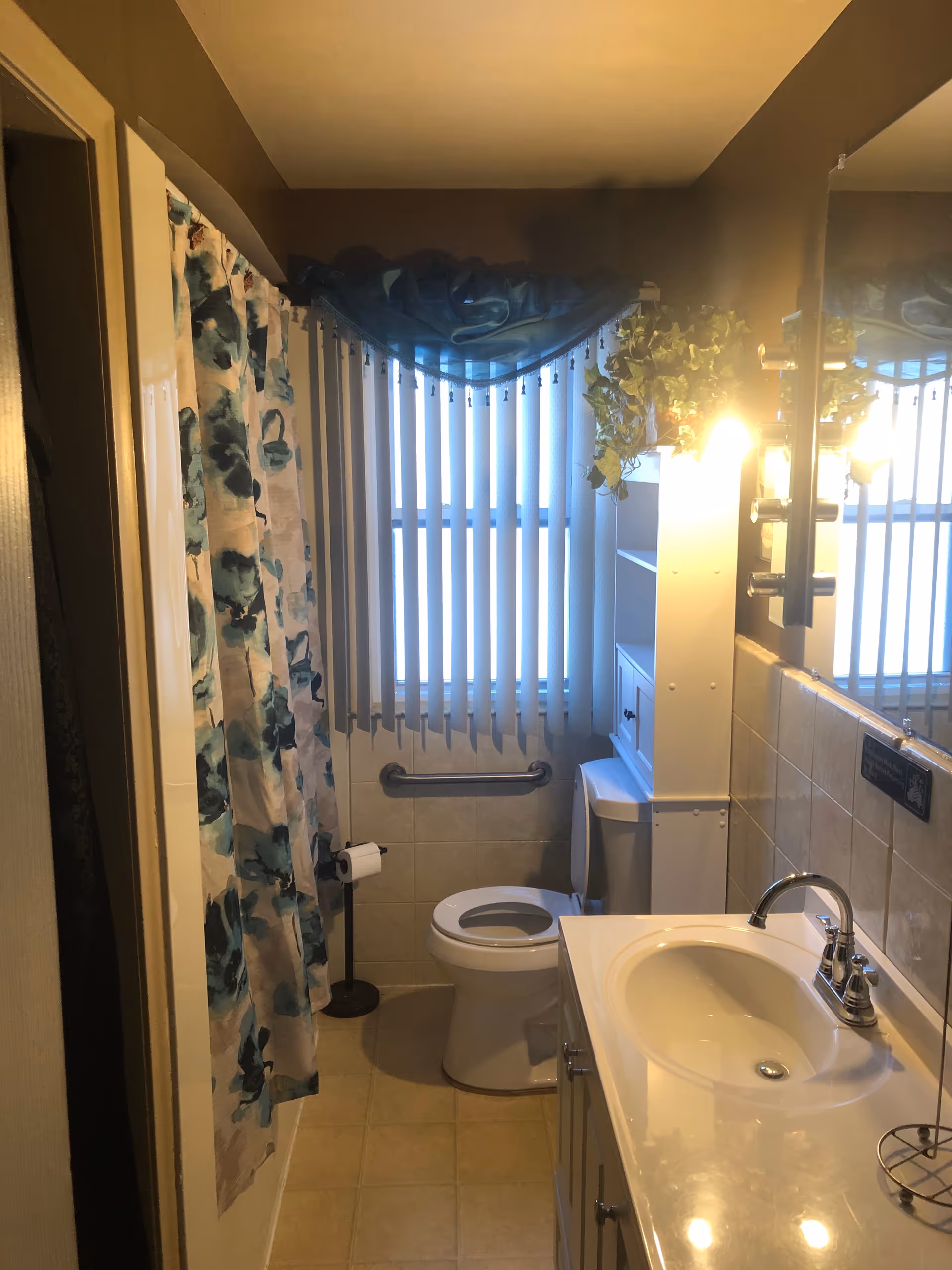 Compact bathroom in a senior living facility featuring toilet, sink, shower curtain, grab bar, blinds.