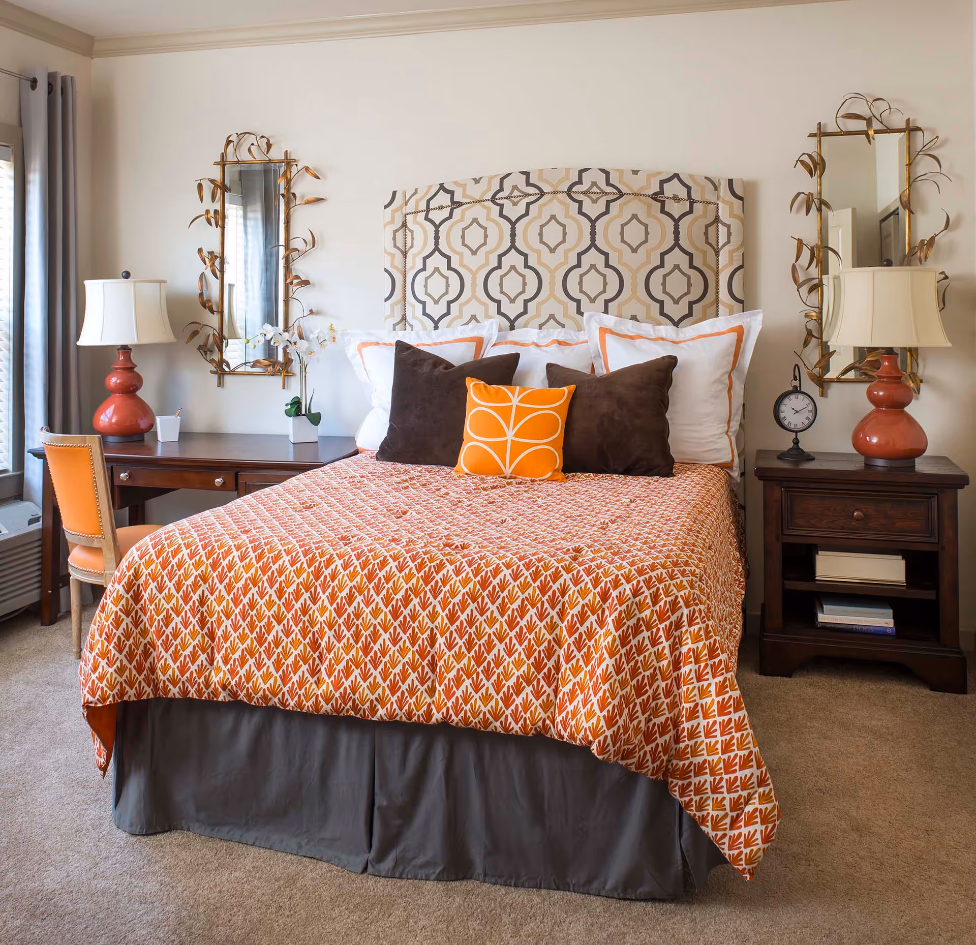 A neatly made bed with a patterned headboard and an orange and white bedspread. The bed has multiple pillows including white, brown, and an orange decorative pillow. On each side of the bed are dark wooden nightstands with matching orange lamps and decorative mirrors above them. There is a chair and desk on the left side near a window with curtains.