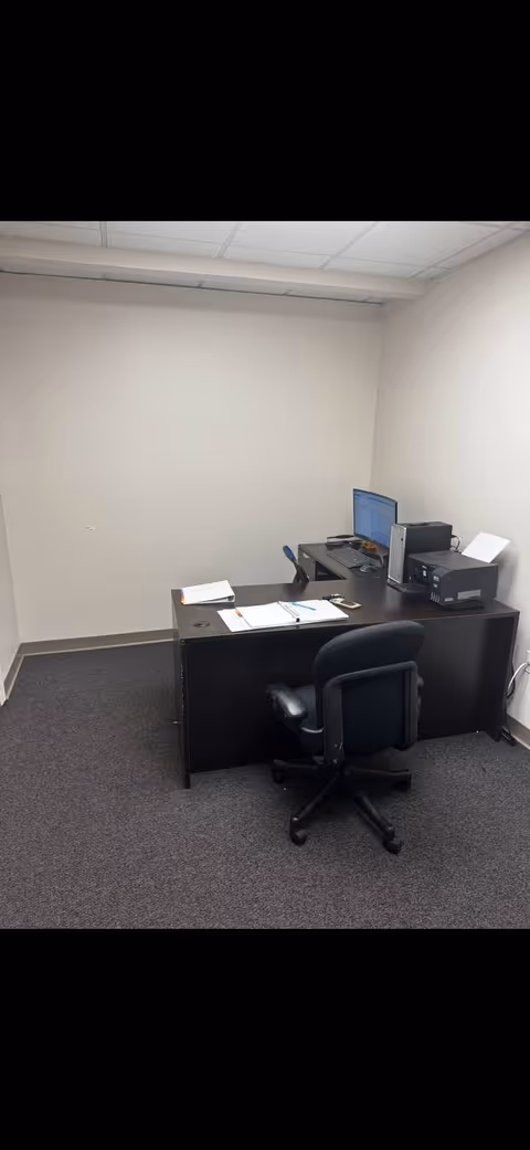 Small office with a desk, chair, computer, and printer in a neutral room.