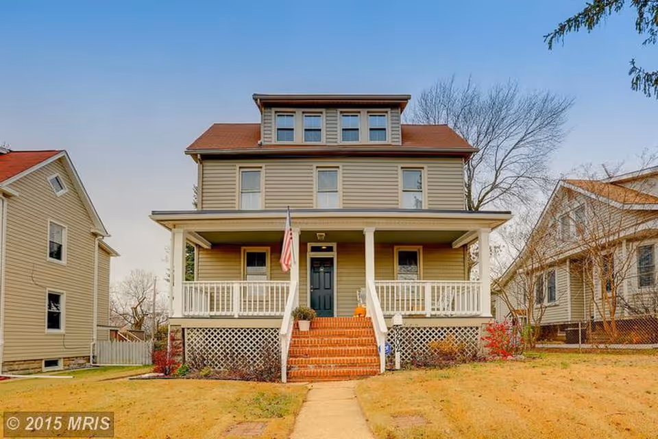 Two-story beige house with a front porch, brick steps, and a flag.