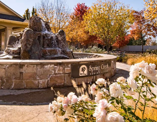 Outdoor scene at Spring Creek Senior Living featuring a stone water fountain with water cascading over rocks, surrounded by blooming white and pink flowers. In the background, there are trees with autumn-colored leaves and a bench along a paved pathway.
