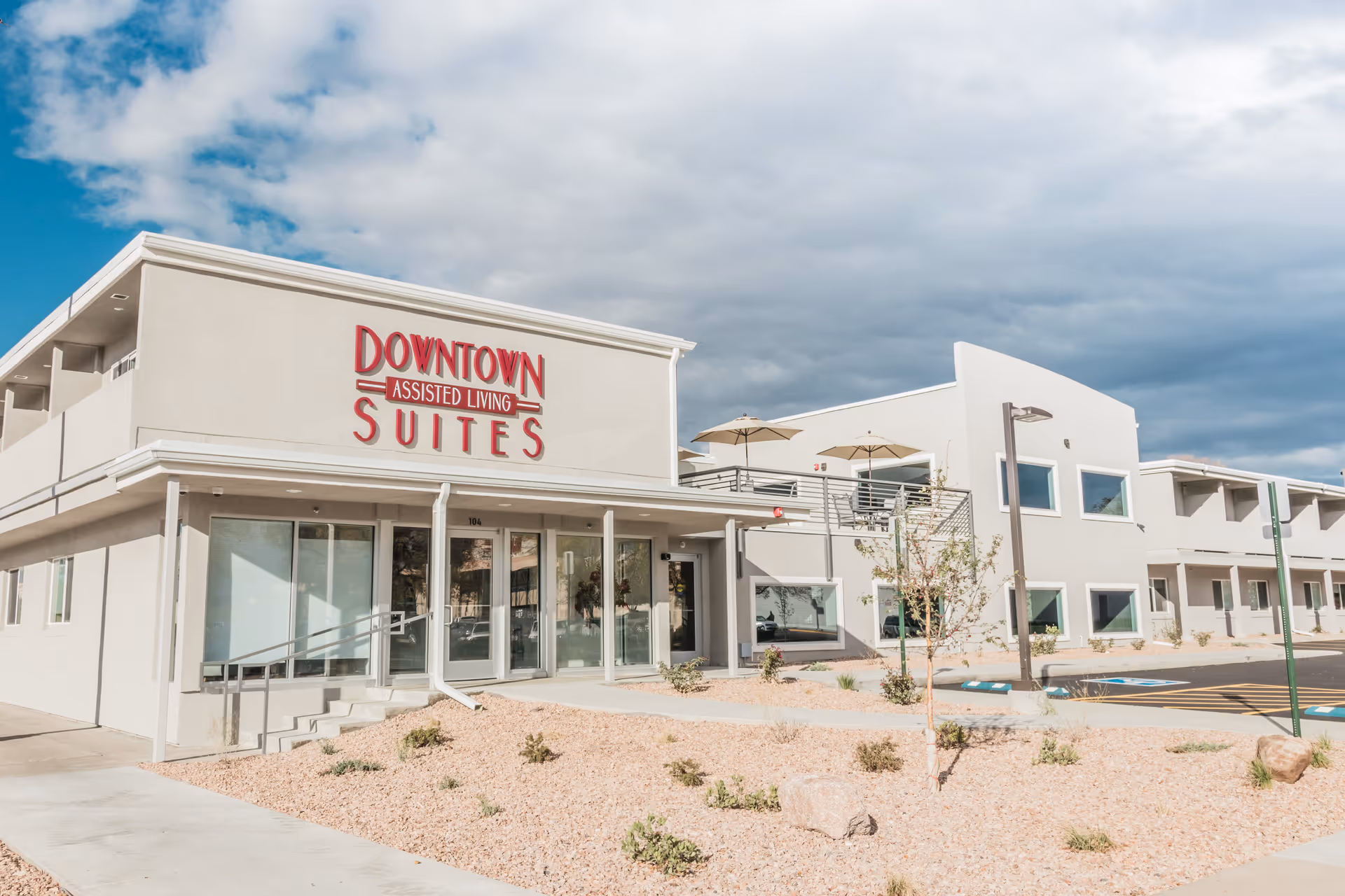 Front exterior of the Downtown Assisted Living Suites building with its sign, entrance, parking area, and desert landscaping under a cloudy sky.