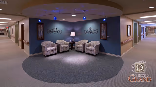 Hallway seating alcove with four upholstered chairs, a side table and lamp under blue pendant lights in a senior living facility corridor.