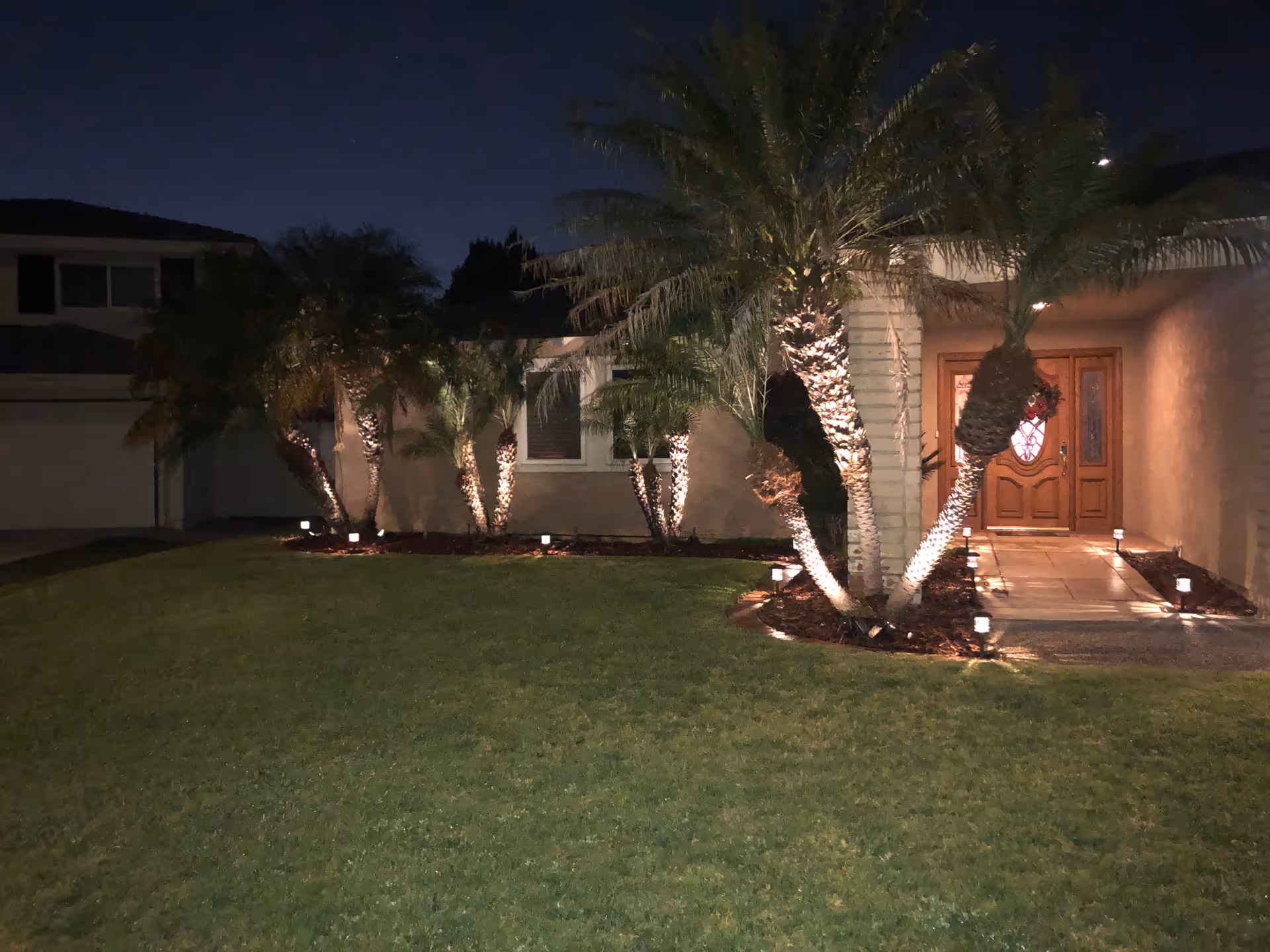 Night view of the front exterior of a house with a well-maintained lawn, palm trees illuminated by ground lights, and a wooden front door with decorative glass panels.