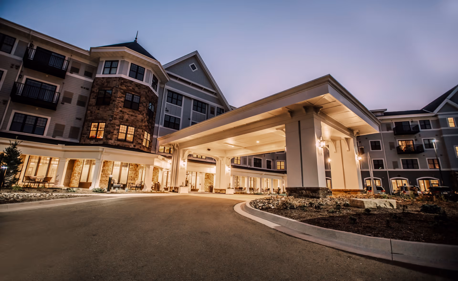 Exterior view of a senior living facility building at dusk with a large covered entrance driveway, stone and siding facade, multiple windows, and outdoor seating areas.