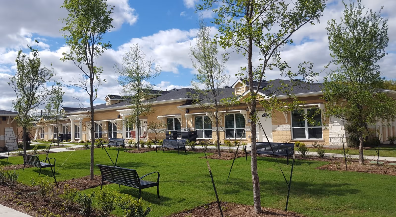 Outdoor view of a senior living facility with a row of single-story beige brick buildings featuring white doors and windows. The foreground shows a well-maintained green lawn with young trees supported by stakes and several black metal benches. The sky is partly cloudy with patches of blue.