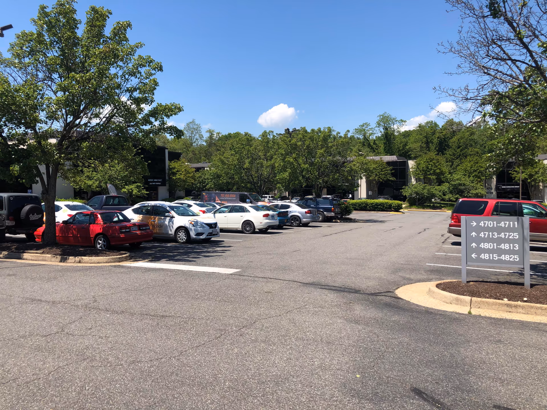 Parking lot in front of a low-rise office building with trees and directional sign.