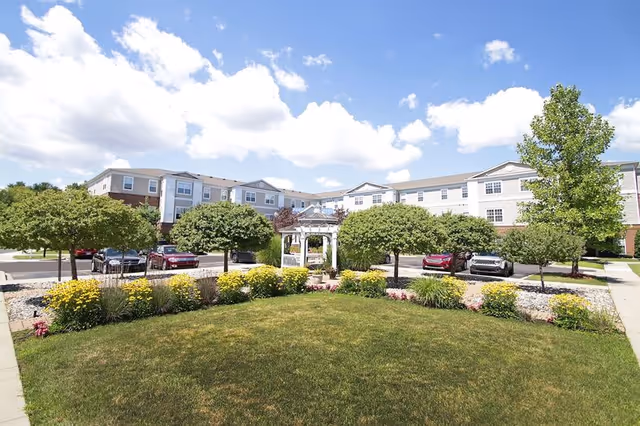 Front exterior of the Lockwood of Clinton senior living building with a landscaped lawn, white gazebo, flowering beds, small trees, and parked cars under a blue sky.