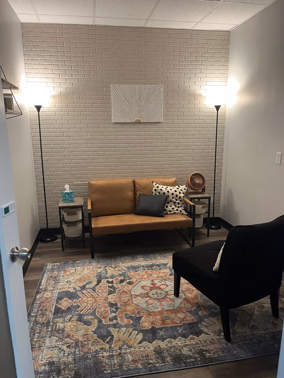 Cozy lounge area with tan sofa, pillows, two floor lamps, and a patterned rug.
