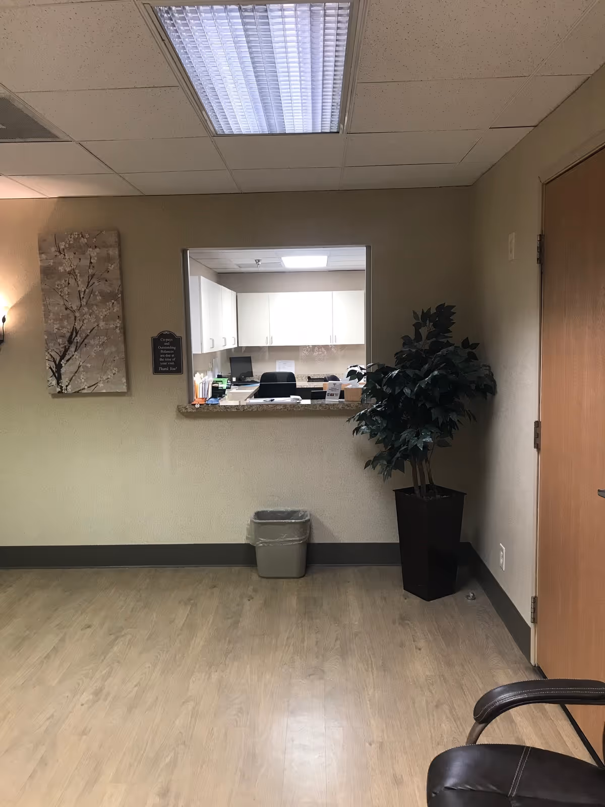 Interior view of a reception window into a small office with a plant and trash can.