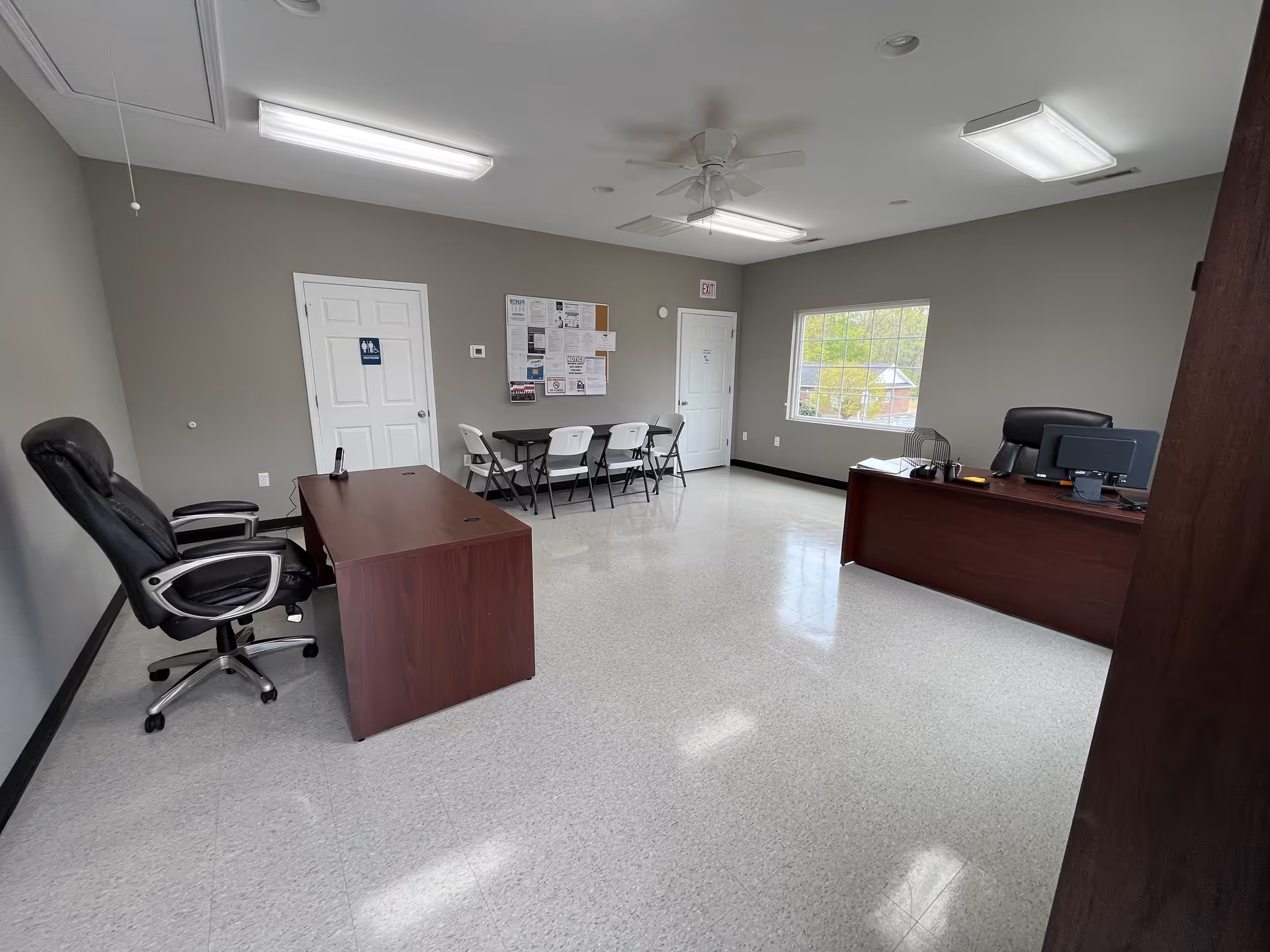 Interior office space with desks, chairs, bulletin board, and a window in grey walls.