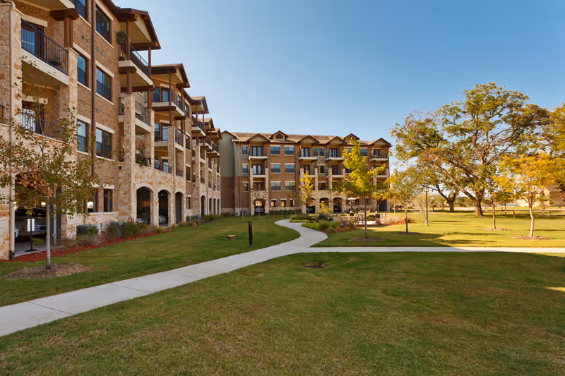 Exterior view of a multi-story brick and stone senior living building with balconies, a winding sidewalk, and lawn with trees under a clear sky.
