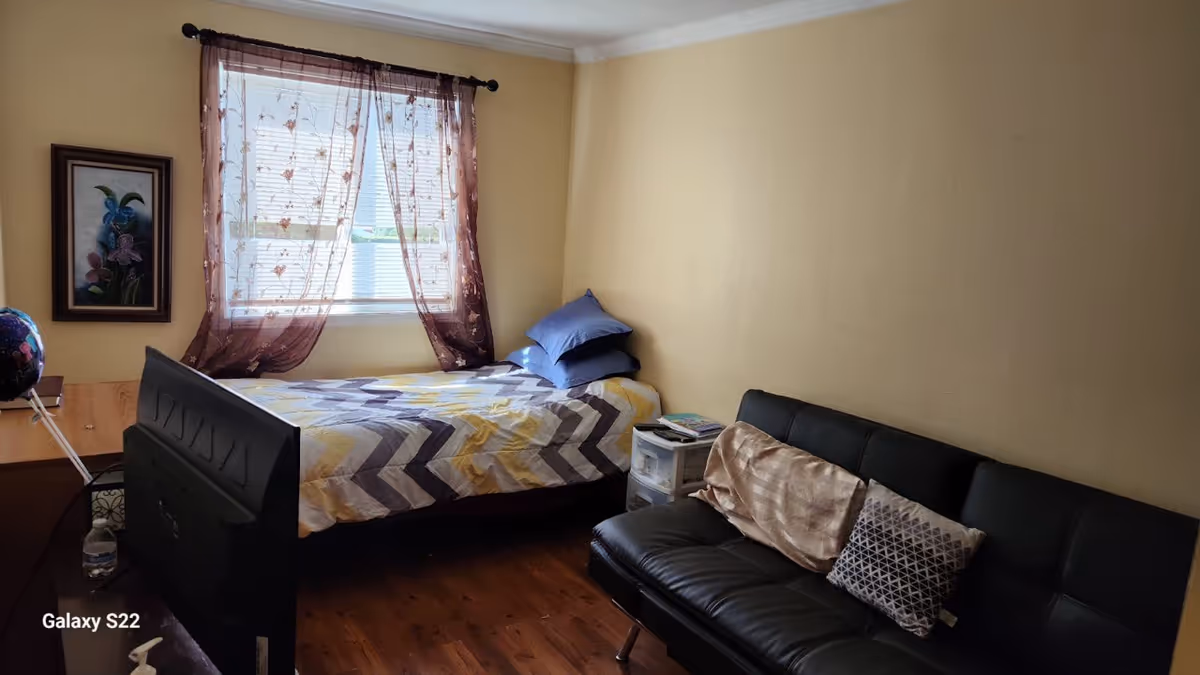 A small bedroom with a bed, black sofa, window with sheer curtains, and wooden floor.