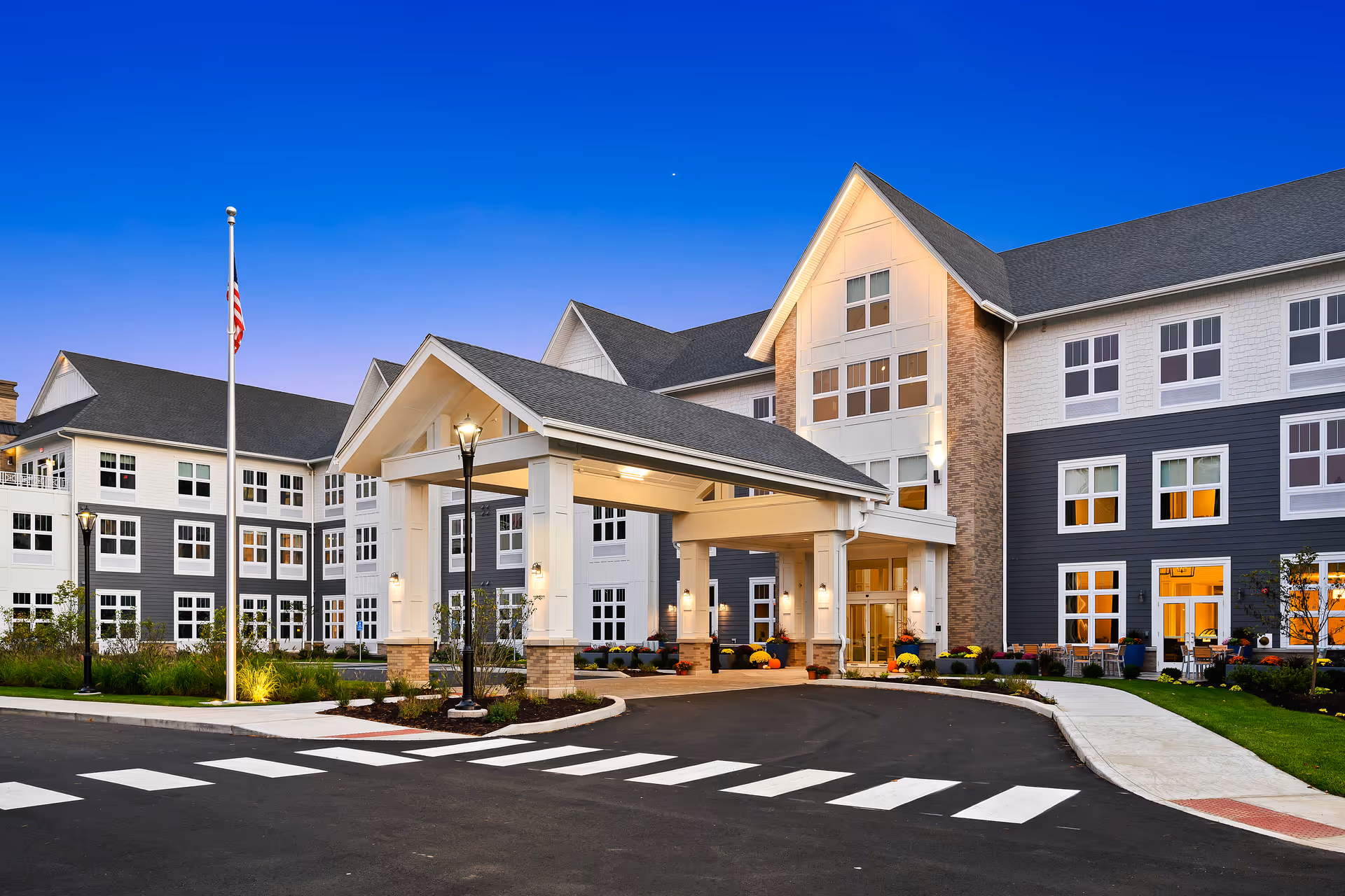 Front entrance of Riverbend at Farmington senior living building with a covered porte-cochere, American flag, and lit windows at dusk.
