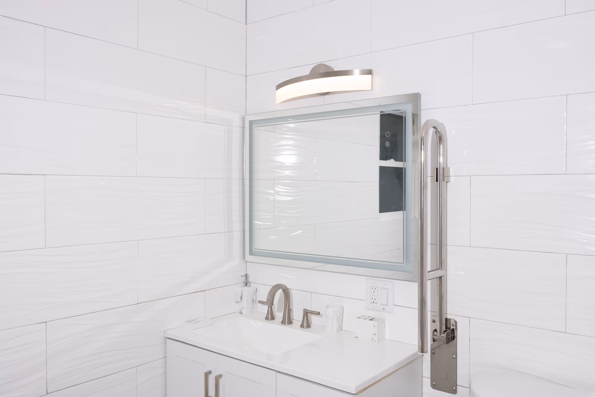 White tiled bathroom with sink, mirror, and wall-mounted accessibility grab bar.