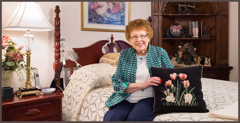 A woman sits on a crocheted-bedspread holding a black floral-embroidered pillow in a traditionally furnished bedroom.