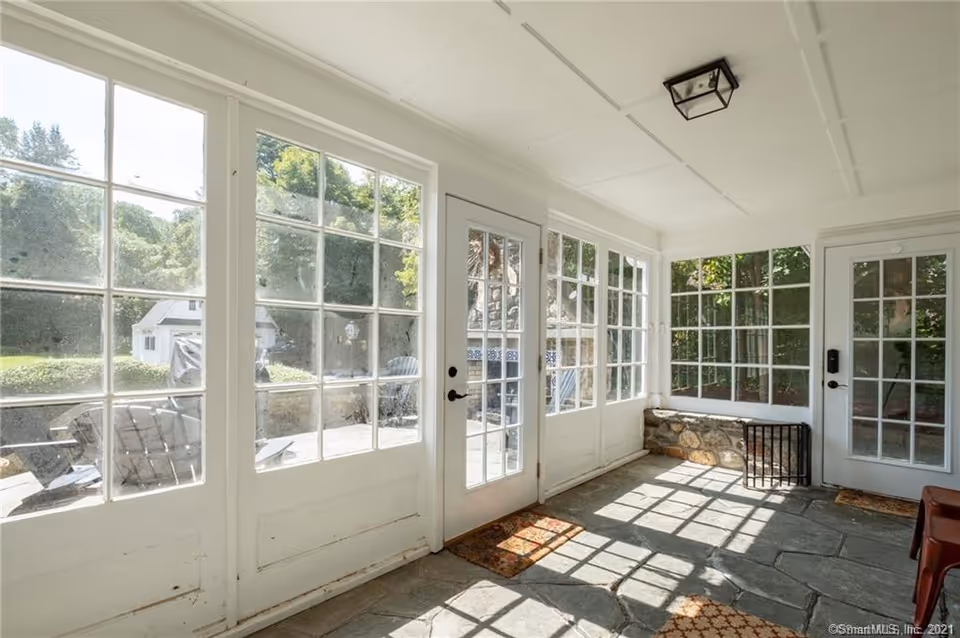 White enclosed porch with large glass doors, stone floor, and garden view outside.