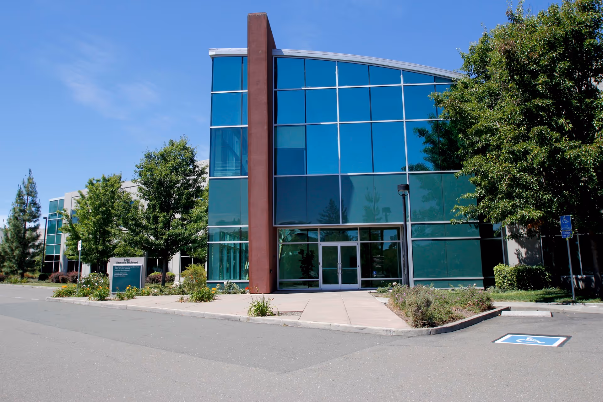 Modern glass-front building with green-tinted windows, trees, and a wheelchair-accessible entrance.