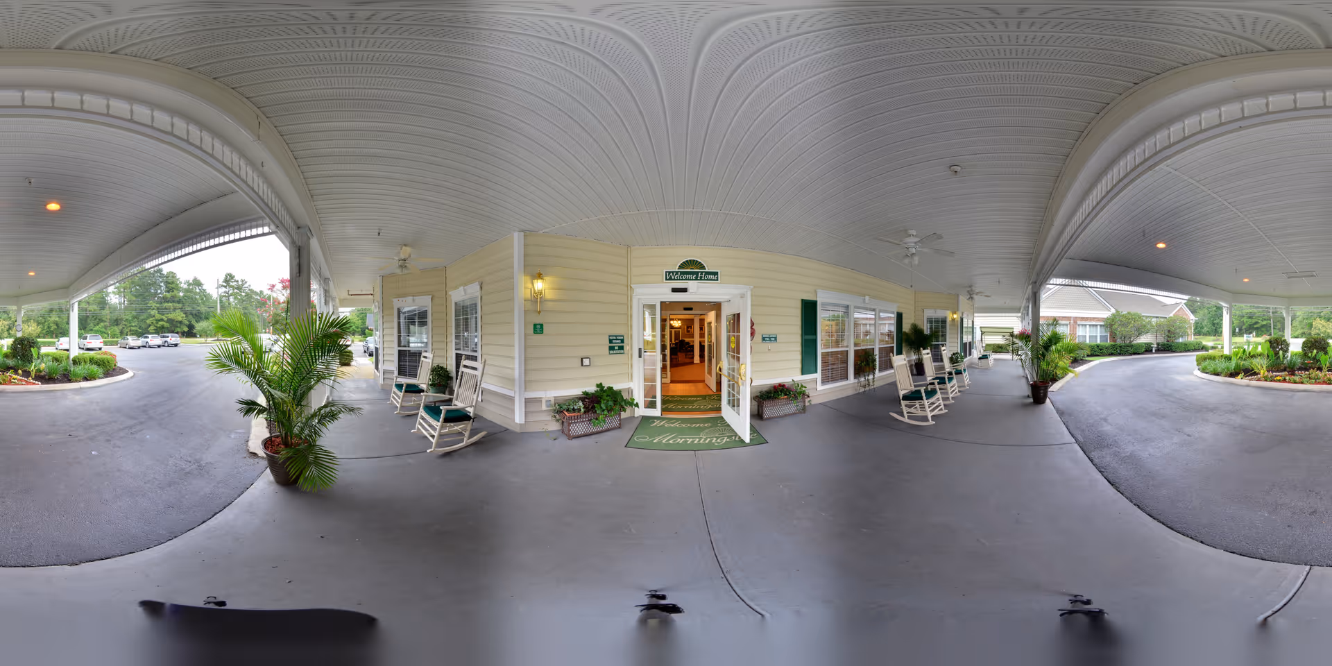 Covered entrance area of The Legacy of Hartsville facility with rocking chairs and potted plants on both sides. The entrance door is open, revealing a welcoming interior. The driveway curves around the entrance with landscaped greenery and parked cars visible in the background.