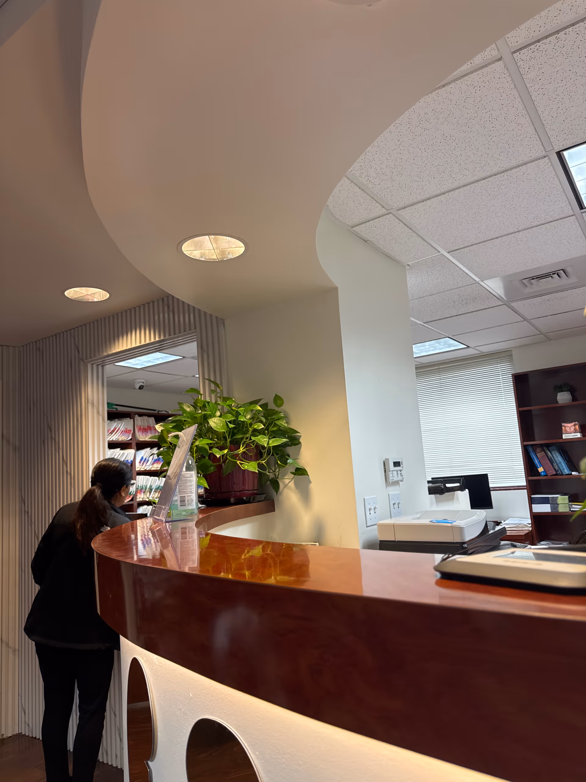 A curved wooden reception desk in a modern office lobby with plants and a staff member.