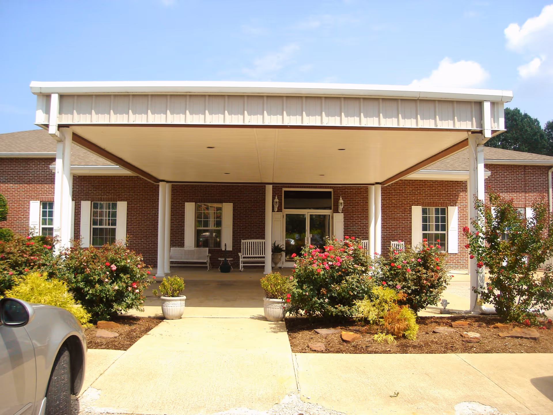 Entrance of a senior living community building with a covered drop-off area supported by white columns. The building has a brick exterior with white window shutters. There are flowering bushes and potted plants along the walkway leading to the entrance. A car is partially visible on the left side of the image.