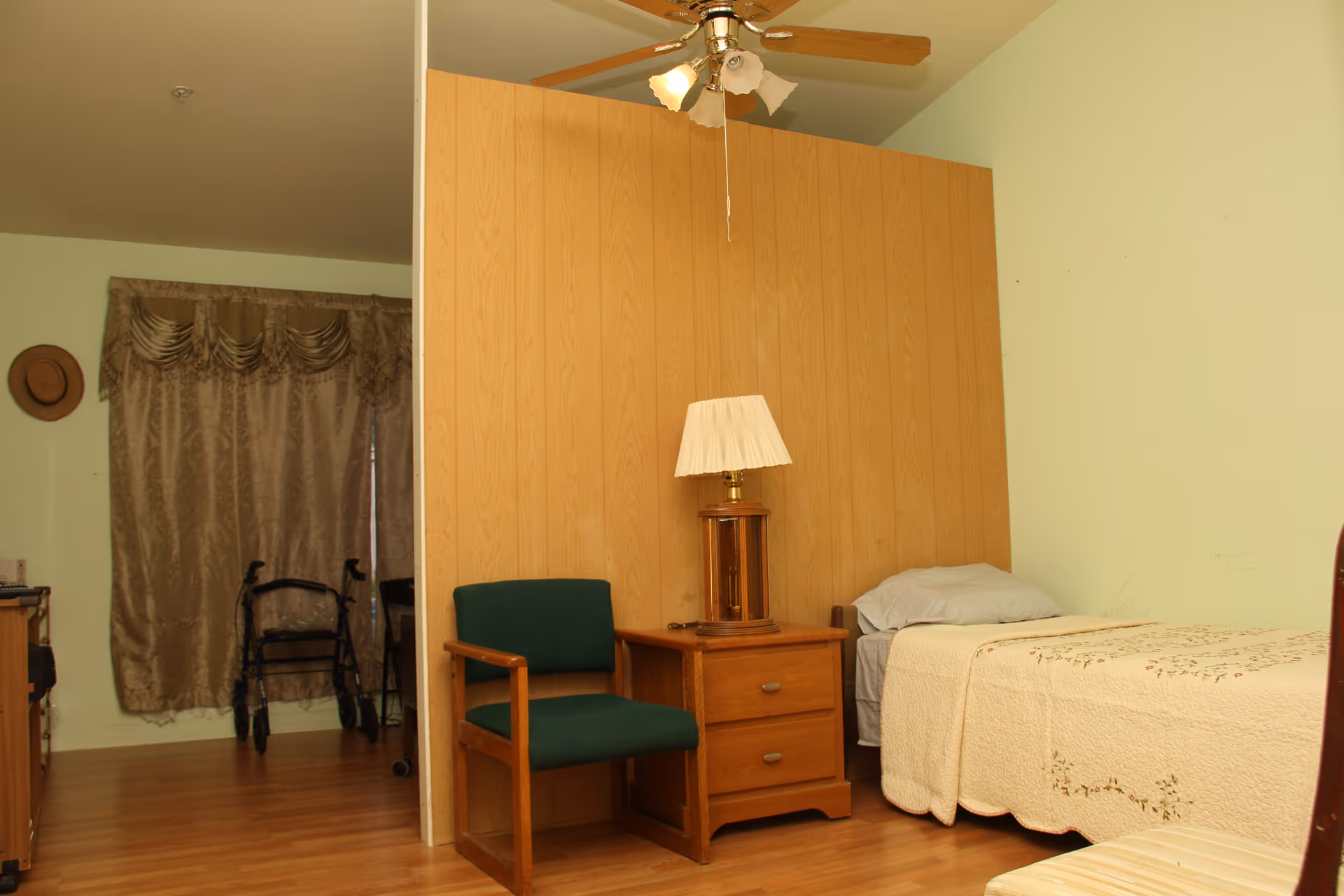 Small bedroom with a single bed, wooden nightstand and lamp, green upholstered chair, and a walker near a curtained window.