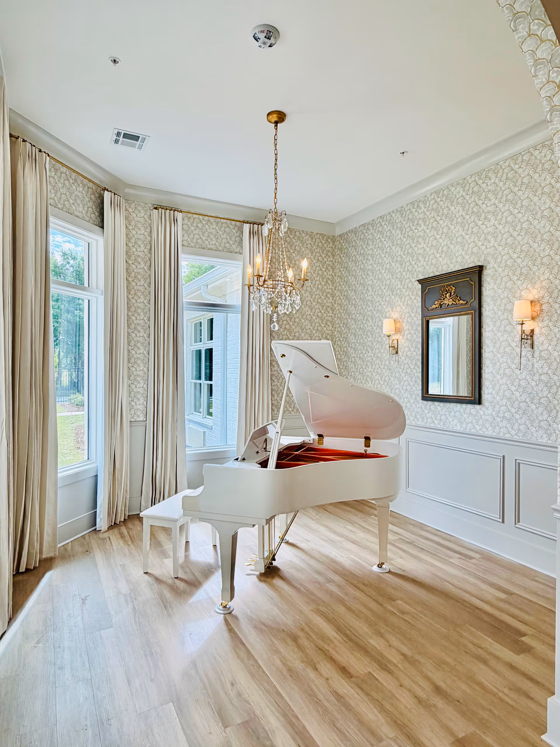 Bright parlor featuring a white grand piano, chandelier, wood floor, and patterned wallpaper.