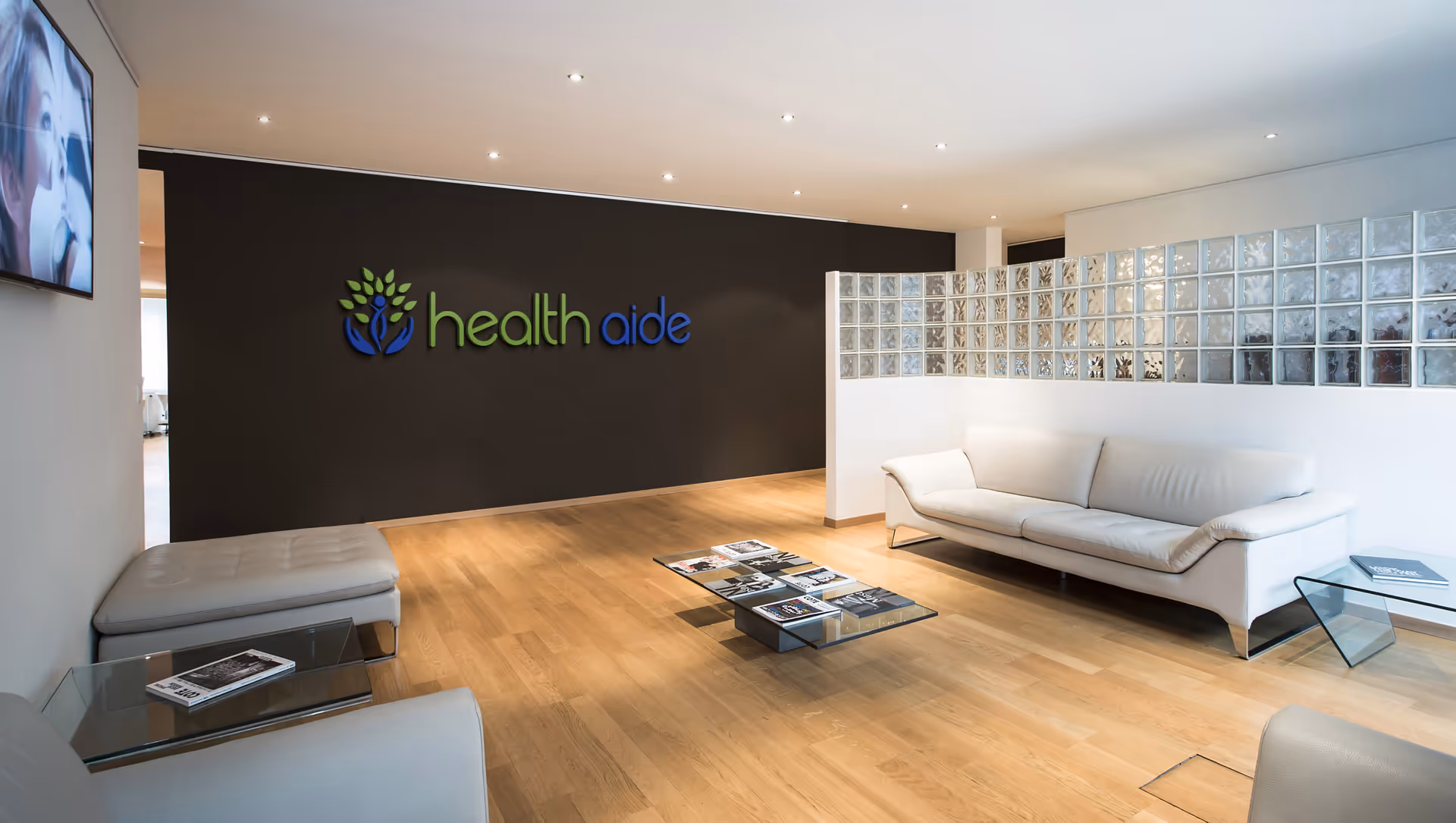Modern reception area with white couches and a dark wall displaying Health Aide logo.
