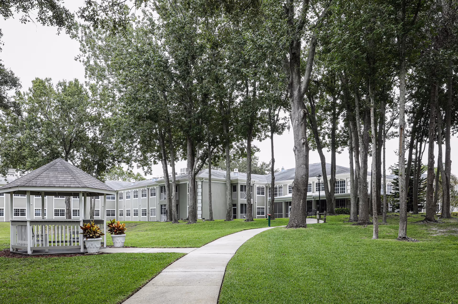 A large two-story senior living building set behind a grassy lawn and trees with a curved sidewalk leading to a gazebo.